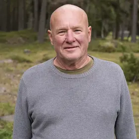En man som heter Crister Boström och är tillsynsperson står på en öppen gräsyta med skog i bakgrunden.