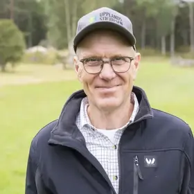 En man som heter Jan Hellberg och är tillsynsperson står framför en stor öppen gräsyta med lövträd i bakgrunden.