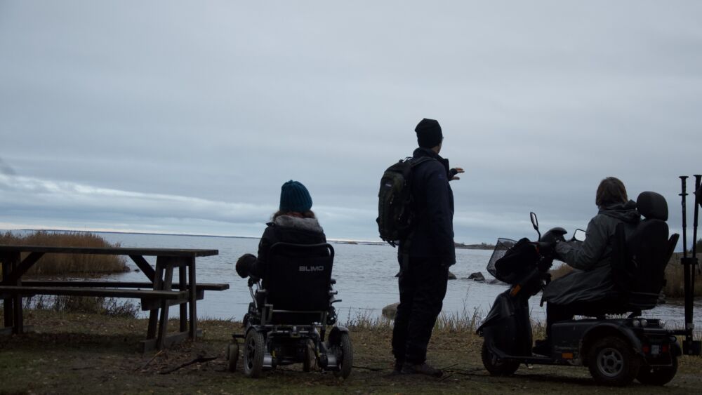 Tre personer ser ut över havet, två av dessa sitter i rullstolar.