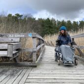 Kvinna i rullstol på träspång med skylt där det står