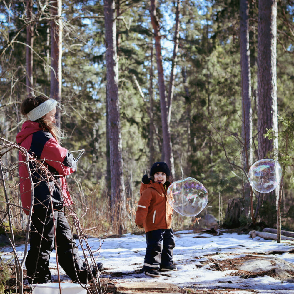 En kvinna och ett barn är i skogen och tittar på stora såpbubblor. Det är snö och de har vinterkläder på sig.