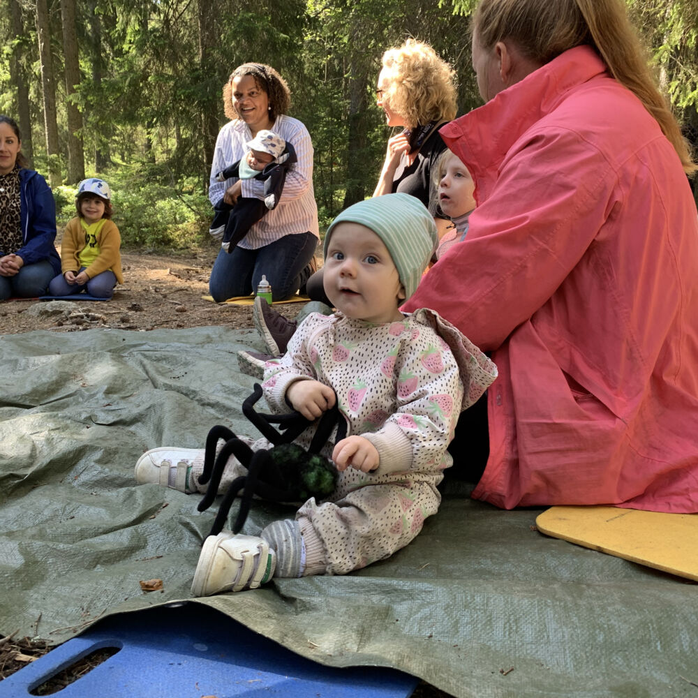 Kvinnor och barn är utomhus och sitter ner. Ett barn med ett mjukisdjur tittar in i kameran.