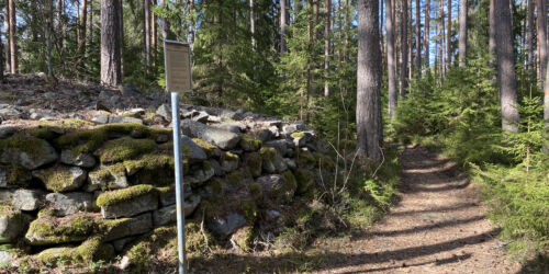 En bred stig leder bort genom en tallskog. Till vänster ses en skylt och bakom den en rund formation bygd av stenar.