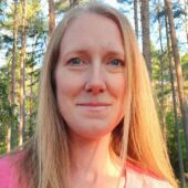 Kvinna med långt blont hår och skog i bakgrunden.