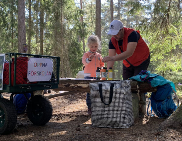 En flicka och en man pysslar med färger ute i skogen. Solen skiner och på sidan står en vagn med texten 