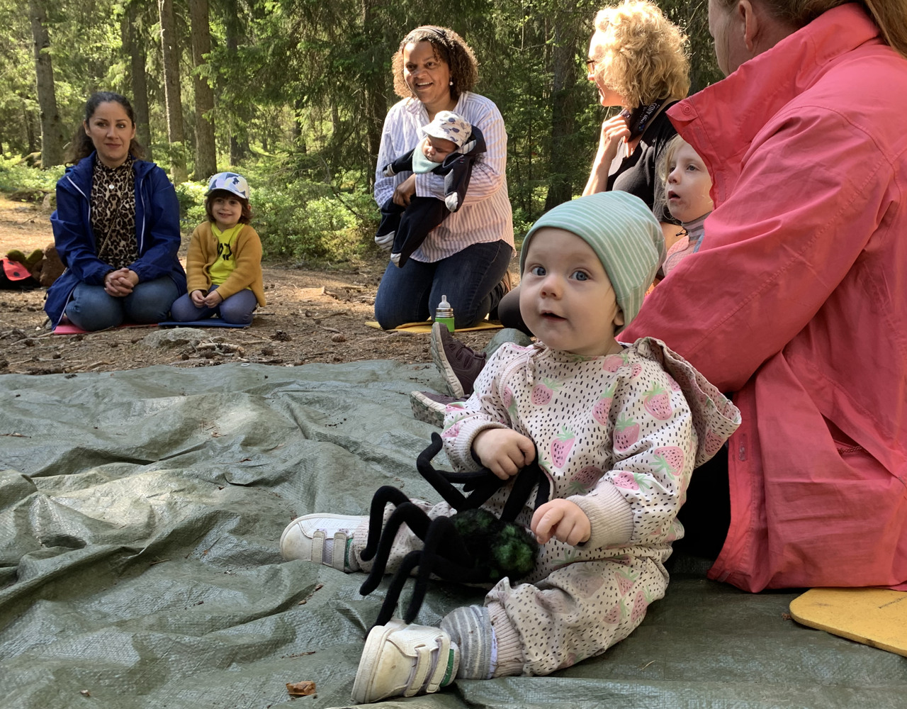 Barn med spindelmjukisdjur sitter i en ring med andra barn och föräldrar utomhus>