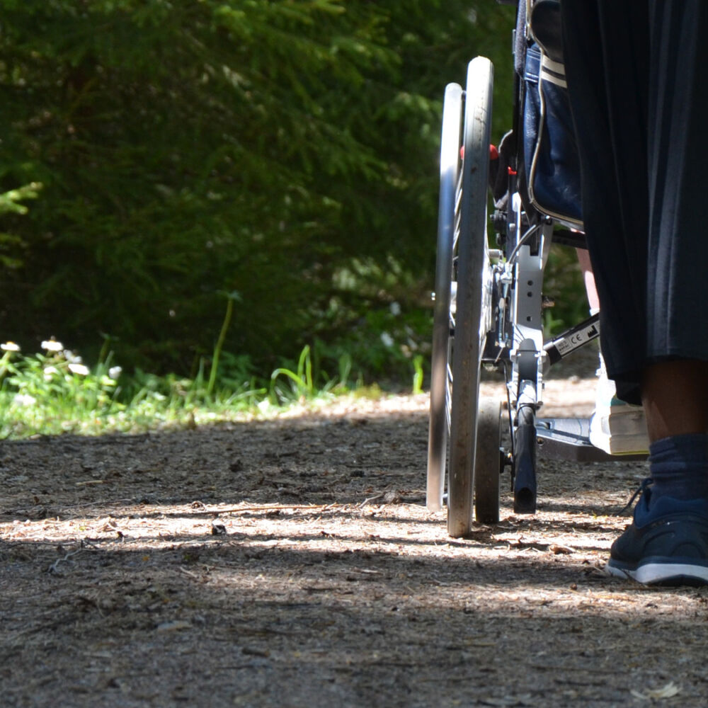 Närbild på fötter, en rullstols hjul och en solbelyst gångväg.