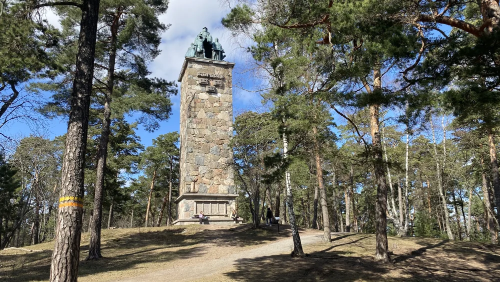 En staty står på en flera meter hög pelare av sten, omgiven av tallar.