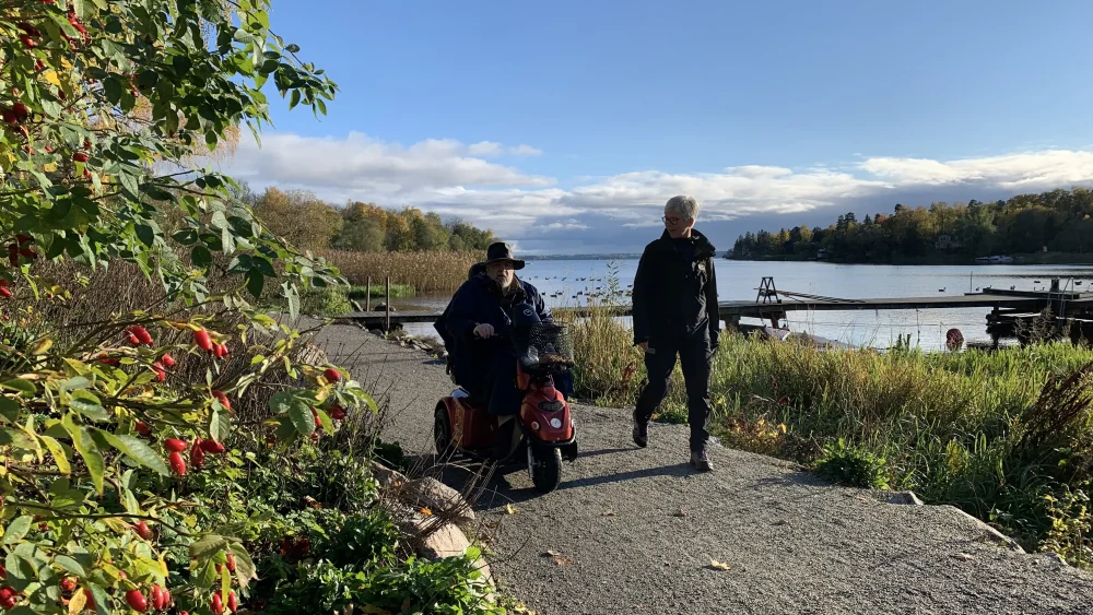 Två personer går på en stig intill ett vattendrag. Den ena personen kör elrullstol.