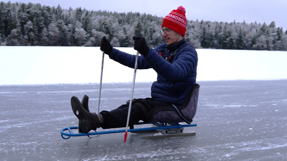 En man åker skridskokälke på isen på en sjö med snö i bakgrunden.