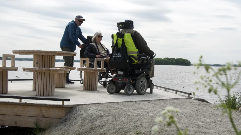 Tre personer är på en brygga vid en sjö. En av dem står och två av dem sitter i varsin rullstol.