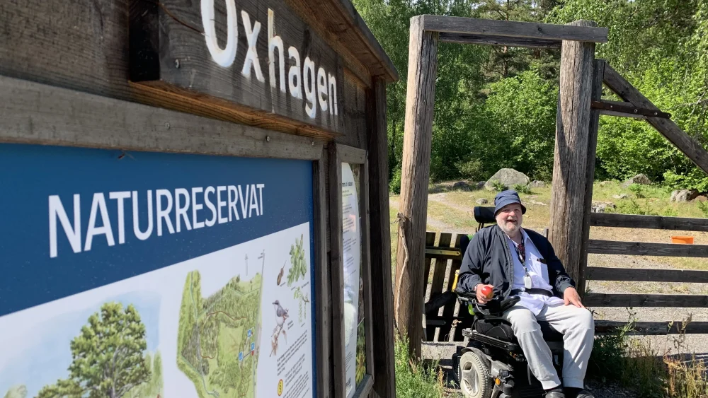 En man i rullstol sitter framför grinden och staketet in till en hage. Bredvid finns en skylt om Oxhagens naturreservat.