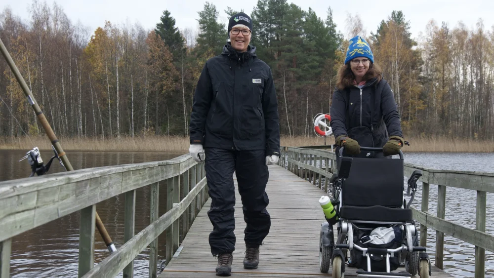 Två kvinnor går på en långa brygga över en sjö, den ena skjuter en rullstol framför sig.