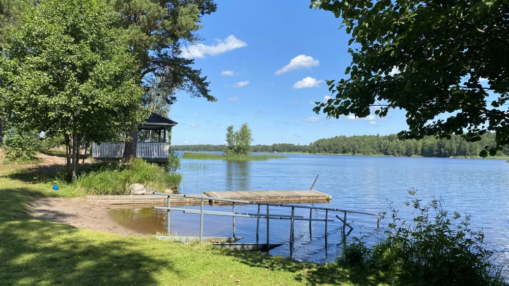 Badplats vid en sjö, med gräsytor, sandstrand, brygga och smal badramp.