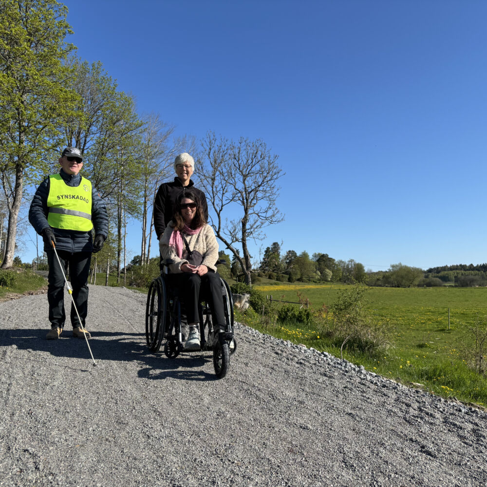 En kvinna i rullstol leds nerför en grusväg.