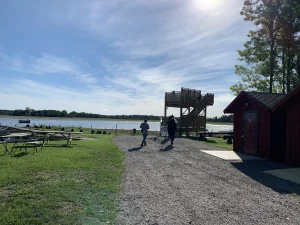 Fågeltorn vid vatten. Ett litet rött hus till höger. Rastbord på gräsyta till vänster i bilden.